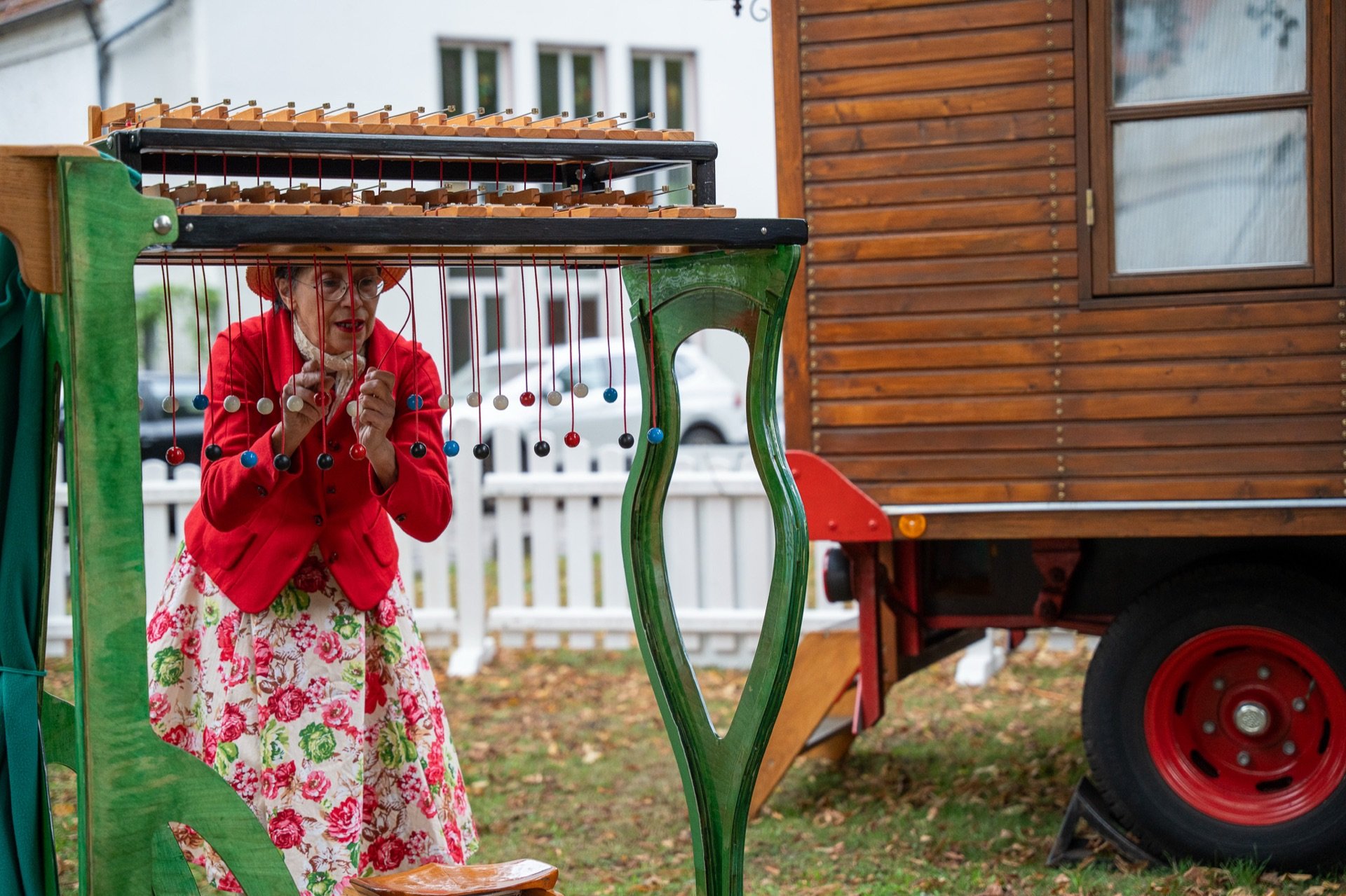 Künstlerin im roten Jackett spielt auf dem Vorplatz an einem Klangspiel neben einem historischen Zirkuswagen