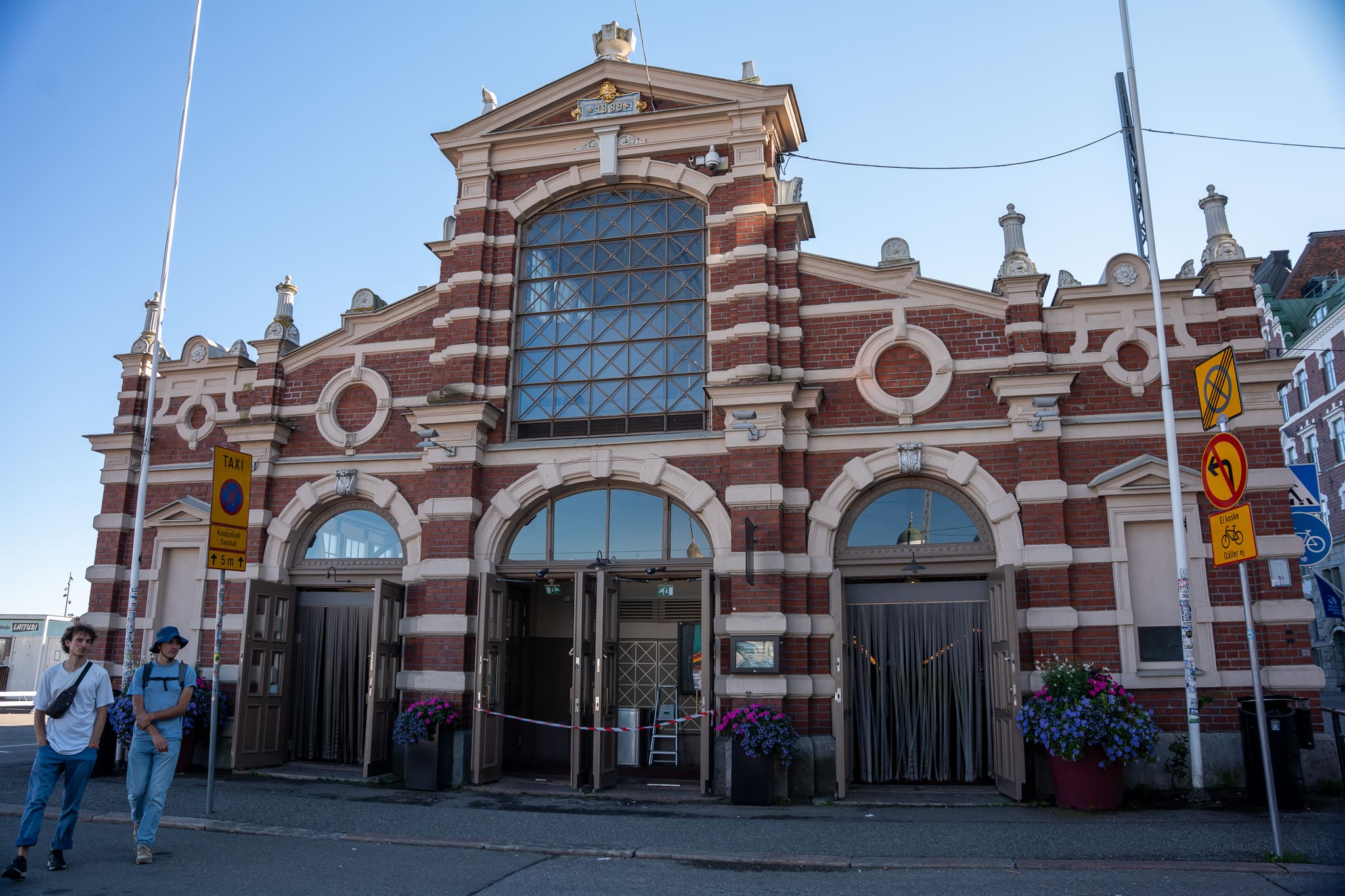 Backsteinfassade der alten Markthalle von Helsinki mit Rundbogenfenstern, davor gehen zwei Passanten vorbei